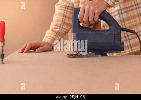 Ein unwiederbringlicher, gesichtsloser, handlicher Mann, der mit einer Puzzle arbeitet. Fokussierter männlicher Holzarbeiter in einer Schutzbrille, der Holzbretter mit einer elektrischen Stichsäge auf einer Werkbank schneidet Stockfoto