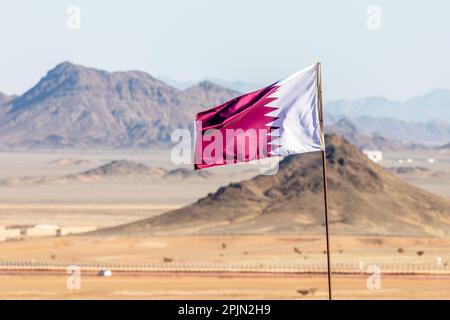 Katar-Flagge, die im Wind winkt, in einer saudiarabischen Wüste mit Bergen im Hintergrund, Al Ula, Saudi-Arabien Stockfoto