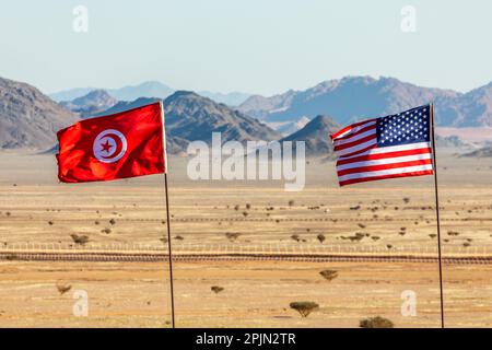 US-amerikanische und tunesische Flaggen, die im Wind miteinander winken, in einer saudiarabischen Wüste mit Bergen im Hintergrund, Al Ula, Saudi-Arabien Stockfoto