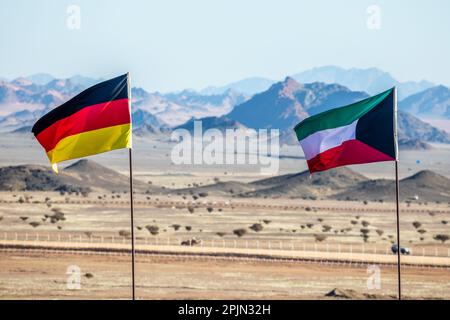 Deutsche und kuwaitische Flaggen, die sich im Wind versammeln, in der saudiarabischen Wüste mit Bergen im Hintergrund, Al Ula, Saudi-Arabien Stockfoto