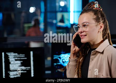 Junge lächelnde Programmiererin, die mit einem Kollegen am Mobiltelefon über das Decodieren von Daten auf Computerbildschirmen in dunklen Büros berät Stockfoto