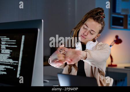 Junge, müde, weibliche IT-Support-Managerin mit Dreadlocks, die sich die Arme vor sich strecken und sich am Arbeitsplatz eine Minute entspannen Stockfoto