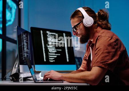 Seitenansicht eines seriösen jungen Mannes mit Kopfhörern, der auf der Laptop-Tastatur tippt, während er am Arbeitsplatz im dunklen Büro sitzt und nachts arbeitet Stockfoto