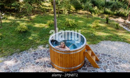 Junge Frau, die in der Holzbadewanne mit Kamin Holz verbrennt und Wasser im Hinterhof in den Bergen erwärmt. High-Angle-Ansicht. Stockfoto