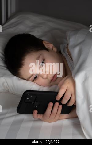 Ein 5-jähriger Junge spielt Spiele auf seinem Handy, während er im Bett liegt. Geräte im Bett vor dem Schlafengehen bei einem Kind Stockfoto