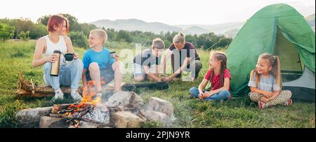 Eine Gruppe lächelnder Kinder unterhalten sich in der Nähe eines rauchigen Lagerfeuers. Sie trinken Tee aus einer Thermoskanne, zwei Brüder richten das grüne Zelt auf. Glückliche Familie Stockfoto