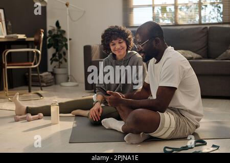 Glückliches, junges interkulturelles Paar, das sich in der Aktivbekleidung ein Online-Sporttraining auf einem Smartphone ansieht, das von einem afroamerikanischen Mann gehalten wird Stockfoto