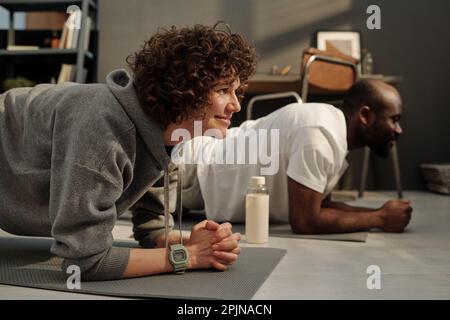 Seitenansicht einer jungen brünetten Frau in grauer Aktivbekleidung und ihres afroamerikanischen Ehemanns, der auf Matten in der Planke steht und beim Workout trainiert Stockfoto