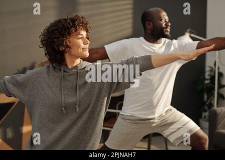 Fröhliche junge brünette Frau in grauem Hoodie, die während des Trainings Arme streckt, während sie vor der Kamera gegen ihren Mann steht Stockfoto