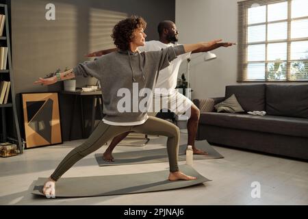 Junge brünette Frau und ihr afroamerikanischer Ehemann stehen auf Matten und machen Dehnübungen während des Trainings im Wohnzimmer Stockfoto