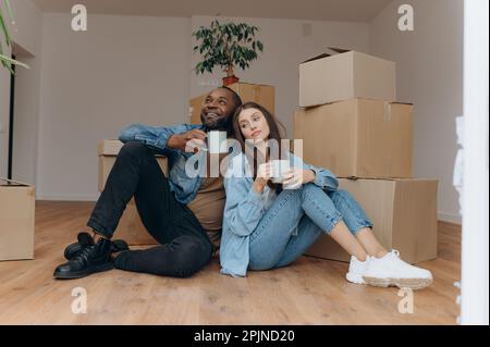 Ein Paar zieht zu Hause ein. Ein gemischtrassiges Paar, das neben Kartons sitzt Stockfoto