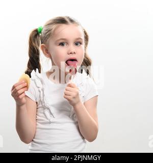 Ein kleines Mädchen leckt einen Lutscher und hält Kartoffelchips in der Hand auf weißem Hintergrund, Babyzähne und Junk-Food. Stockfoto