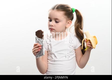 Schokolade und Banane, Auswahl zwischen gesunden und leckeren, gesunden und kalorienreichen Speisen, kleines Mädchen wählt Schokolade und hält Banane, Kopierraum und Stockfoto