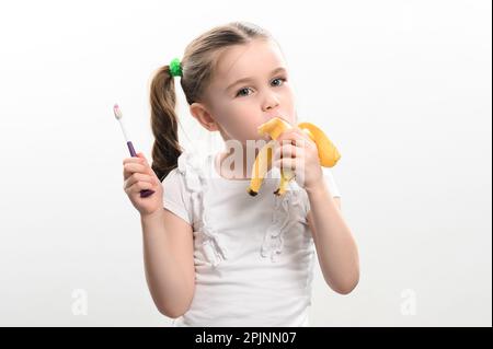 Ein kleines Mädchen isst eine Banane und hält eine Zahnbürste in der Hand, Studiofoto auf weißem Hintergrund, eine Banane hilft gegen Karies, eine Idee für Zahnärzte Stockfoto