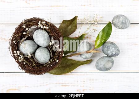 Braunes Zweignest mit drei grauen Ostereiern, Federn, getrockneten Magnolienblättern und Zweigen von Zigeunern auf Holzhintergrund. Minimalistisches Osterfest Stockfoto