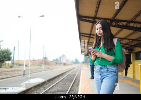Hispanische junge Frau, die auf ihrem Handy in sozialen Medien surft, in einem Bahnhof. Speicherplatz kopieren Stockfoto