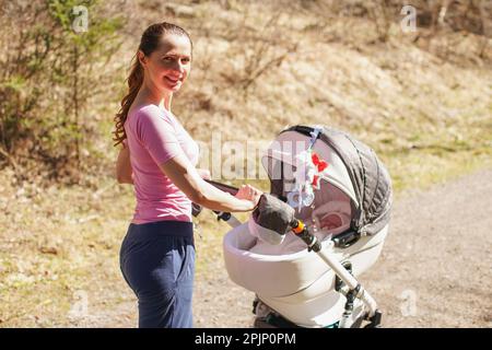 Junge Frau trägt lässige Kleidung und schiebt Kinderwagen auf Asphaltstraße, sonniger Tag, Wald im Schatten Stockfoto