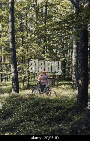 Aktive Frau, die freie Sommerferienzeit auf einer Fahrradtour in einem Wald verbringt. Frau trägt Fahrradhelm und Handschuhe halten Fahrrad mit Korb Stockfoto