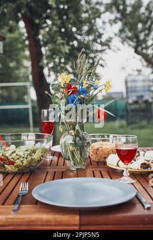 Abendessen in einem Apfelgarten auf Holztisch mit Salaten und Wein mit Blumen dekoriert. Nahaufnahme des Tisches mit Essen für Familienessen vorbereitet Stockfoto