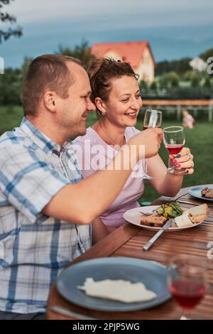Familie, die während des Sommers ein Picknick im Freien in einem eigenen Garten toast. Nahaufnahme von Leuten, die Weingläser mit Rotwein halten Stockfoto