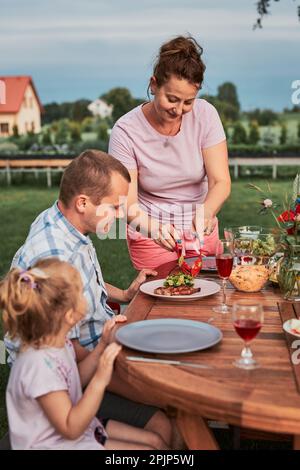 Familie mit einer Mahlzeit vom Grill während des Sommer Picknick im Freien Abendessen in einem Hausgarten. Nahaufnahme von Leuten, die an einem Tisch mit Essen und Geschirr sitzen Stockfoto