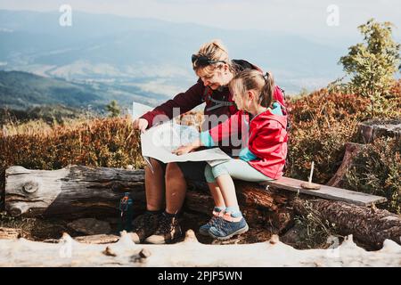 Familienausflug in die Berge. Mutter und ihre kleine Tochter untersuchten eine Karte, saßen auf dem Stumpf, machten während der Sommerreise Pause Stockfoto