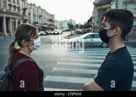 Mann und Frau sprechen und warten an der Fußgängerüberquerung im Stadtzentrum tragen die Gesichtsmasken, um Virusinfektion zu vermeiden und die Ausbreitung zu verhindern Stockfoto