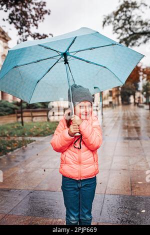 Kleines lächelndes glückliches Mädchen hält großen blauen Regenschirm während des Spaziergangs An regnerisch düsteren Herbsttag Stockfoto