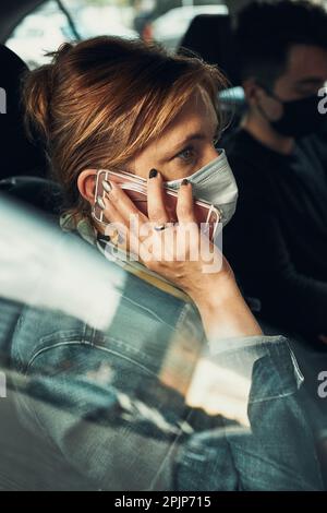 Frau, die mit dem Smartphone telefoniert und in einem Auto sitzt, trägt die Gesichtsmaske, um eine Virusinfektion zu vermeiden und die Ausbreitung der Krankheit rechtzeitig zu verhindern Stockfoto
