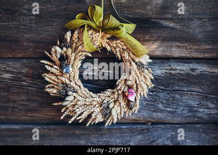 Kranz aus goldenen Ähren aus Weizen, getrockneten Blumen und Kräutern, gebunden mit rotem Band, an der Holzwand aufgehängt. Natürliche rustikale Dekoration Stockfoto