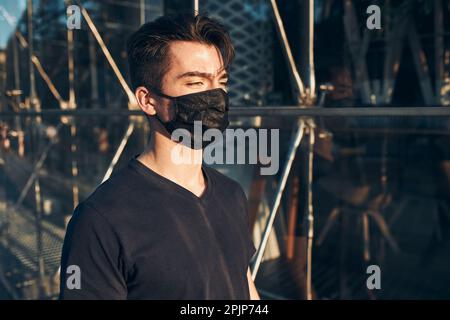 Junger Mann, der abends im Stadtzentrum entlang der Glaswarenfront spazierend, wegschaut, die Gesichtsmaske trägt, um Virusinfektionen zu vermeiden und sich zu verjüngt Stockfoto