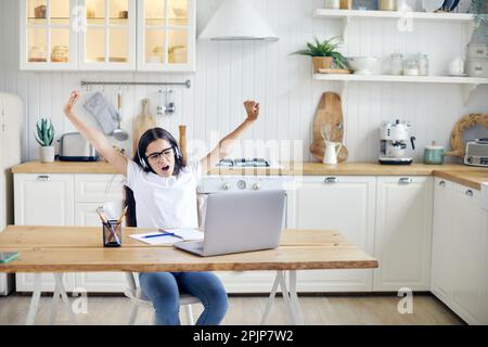 Hübsches 12s-jähriges Mädchen in Kopfhörern sitzt am Tisch mit Laptop, beendet Online-Kurs, Unterricht mit Lehrer, macht Aufgaben, macht Pause, entspannt mit Armen Stockfoto