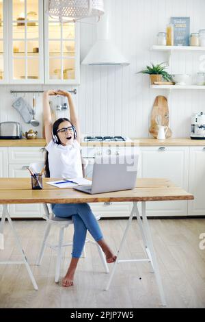 Hübsches 12s-jähriges Mädchen in Kopfhörern sitzt am Tisch mit Laptop, beendet Online-Kurs, Unterricht mit Lehrer, macht Aufgaben, macht Pause, entspannt mit Armen Stockfoto