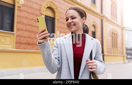 Eine elegante junge Frau, die einen Mantel trägt und auf ihrem Handy Nachrichten liest, während sie die Straße entlang läuft und lächelt. Stockfoto