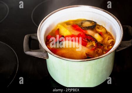 Kochende und sprudelnde Suppe aus rotem und gelbem Pfeffer und Tomate in grünem Topf auf schwarzem Keramikkochfeld. Zubereitung der Suppe aus Pfeffer und Tomate. Stockfoto