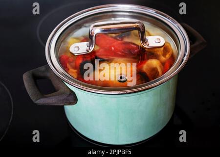 Suppe aus Pfeffer, Zwiebel und Tomate in grünem Topf auf Keramikkochfeld. Stockfoto