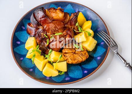 Gebacken bis goldbraun gewürzter Schweinebauch, Knoblauch und Kartoffel auf dekorativem blauem Teller, Gabel auf weißem Hintergrund. Stockfoto