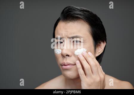 Stirnrunzeln-Mann, der Feuchtigkeitscreme oder Sonnencreme auf das Gesicht aufträgt Stockfoto