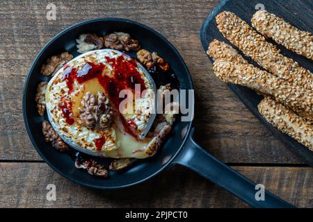 Blick auf gebackene Bratpfanne, Camembertkäse, Walnüsse, Honig, Marmelade und Brotsticks mit Sesamsamen auf Holztisch-Hintergrund, Nahaufnahme, Draufsicht Stockfoto