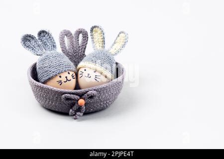 Zwei Ostereier mit lustigen Gesichtern und graue Häkelhüte mit Hasenohren in einem dekorativen Korb auf grauem Hintergrund. Frohes Osterkonzept. Begrüßung C. Stockfoto