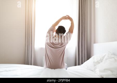 Rückansicht eines Mannes im Pyjama, der auf dem Bett sitzt und sich streckt Stockfoto