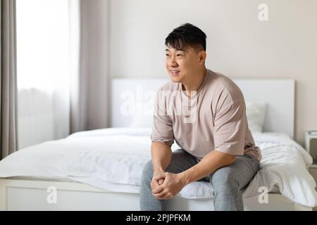 Ein glücklicher asiatischer Mann, der einen glücklichen Morgen genießt, der auf dem Bett sitzt Stockfoto