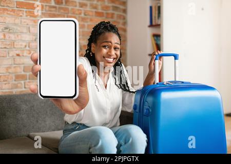 Glückliche schwarze Touristenfrau, die ein Handy mit leerem Bildschirm vor der Kamera zeigt, sitzt zu Hause in der Nähe des gepackten Koffers, Modell Stockfoto