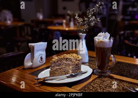 Ein Stück Lviv Marzipan-Kuchen auf einer weißen Keramikplatte, Aareto-Kaffee in einem transparenten Glas, Serviettenhalter auf einem Restauranttisch mit verschwommenem Backgro Stockfoto