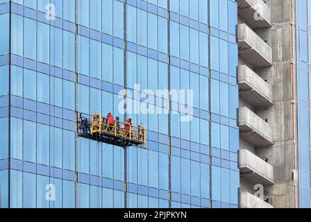 Bauarbeiter auf einer Hängebrücke montieren die Glasfassade eines mehrstöckigen Wolkenkratzers. Stockfoto