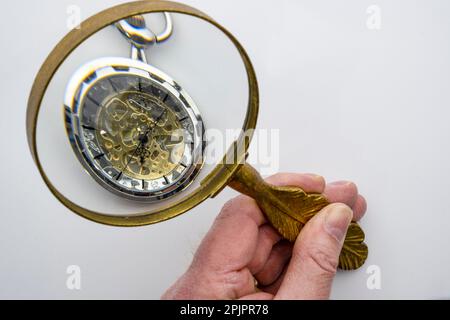 Eine Hand mit einer Lupe, die eine Taschenuhr mit sichtbaren Zahnrädern untersucht, isoliert auf weißem Hintergrund, Konzeptzeitmanagement Stockfoto