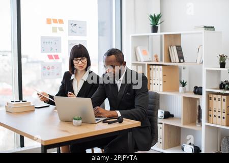 Fokussierter afrikanischer Geschäftsmann und sein netter weißer Assistent analysieren Daten auf einem modernen Laptop, während er an einem kreativen Arbeitsplatz sitzt. Geschäftskollegen diskutieren im Büro über ein neues Finanzprojekt. Stockfoto