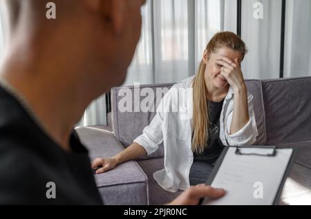 Ein männlicher Psychologe konsultiert eine Patientin, macht Notizen. Eine Frau spricht emotional über sich selbst, bedeckt ihr Gesicht mit ihren Händen. Stockfoto