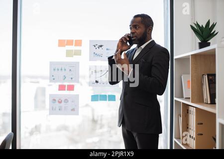 Ein afroamerikanischer Mann im Business-Anzug telefoniert mit einem modernen Smartphone in einem komfortablen Bürozimmer. Chefadministrator interagiert mit anderen Führungskräften über drahtlose Technologie. Stockfoto