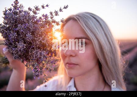 Nahaufnahme einer jungen Frau mit einem Strauß lila Lavendel vor dem Gesicht. Blondes Jahrtausendmädchen mit sauberer Haut ohne Make-up in Su-Strahlen Stockfoto